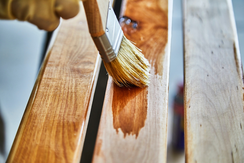 Man's hand holding a paint brush applying varnish to birch wood slats with shallow depth of field